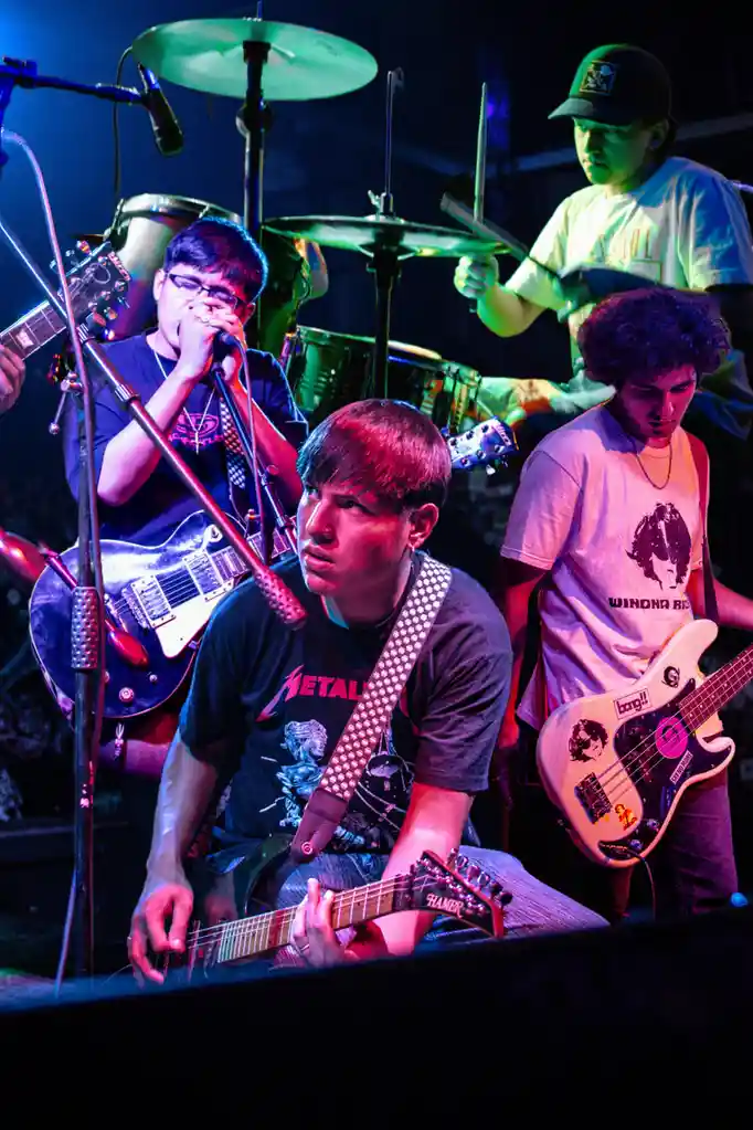 A photo of a four-member rock band performing live on stage under colorful stage lights. The lead guitarist in the…