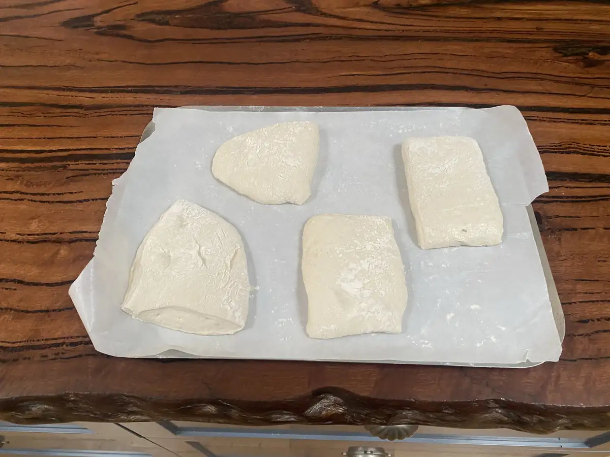 Four irregularly shaped, flour-dusted dough pieces sit on parchment paper atop a baking tray, placed on a dark wooden…