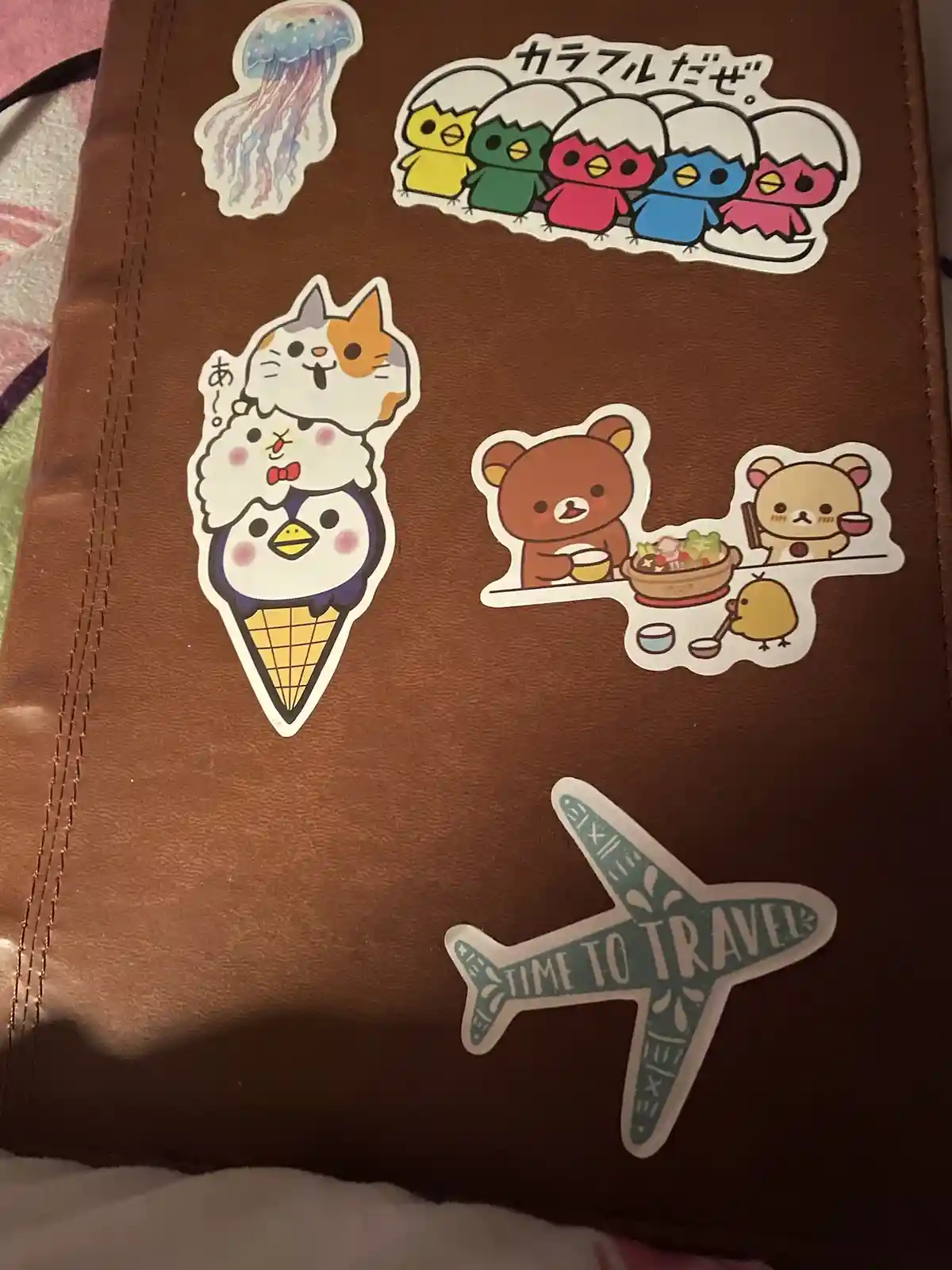A brown leather-bound notebook or journal is covered with various cute Japanese-style stickers. Visible stickers include a…