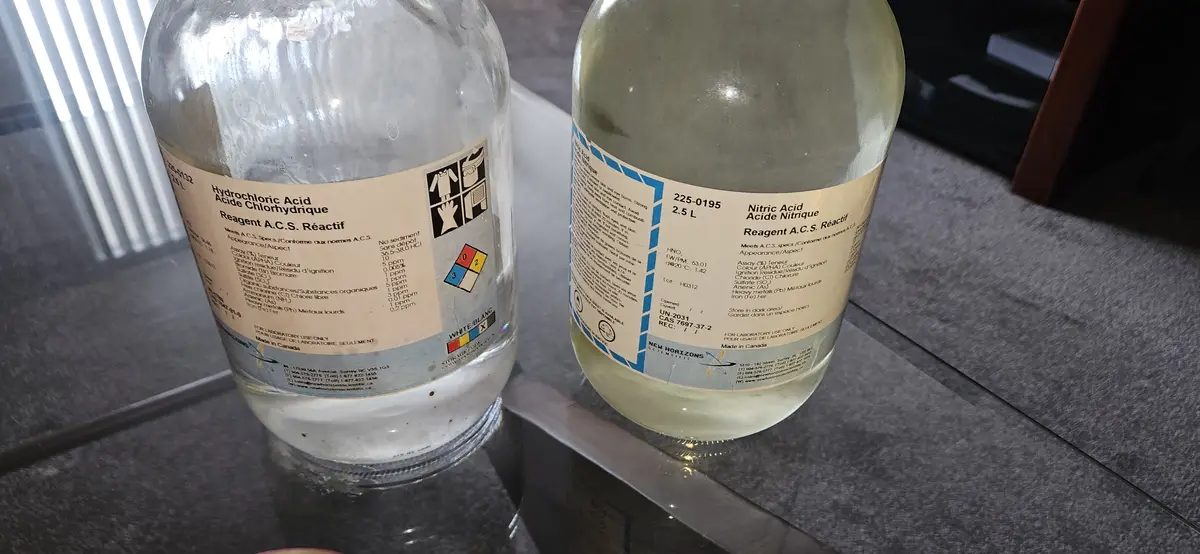 Two large glass bottles labeled 'Hydrochloric Acid' and 'Nitric Acid' sit on a glass table, both marked 'Reagent A.C.S…