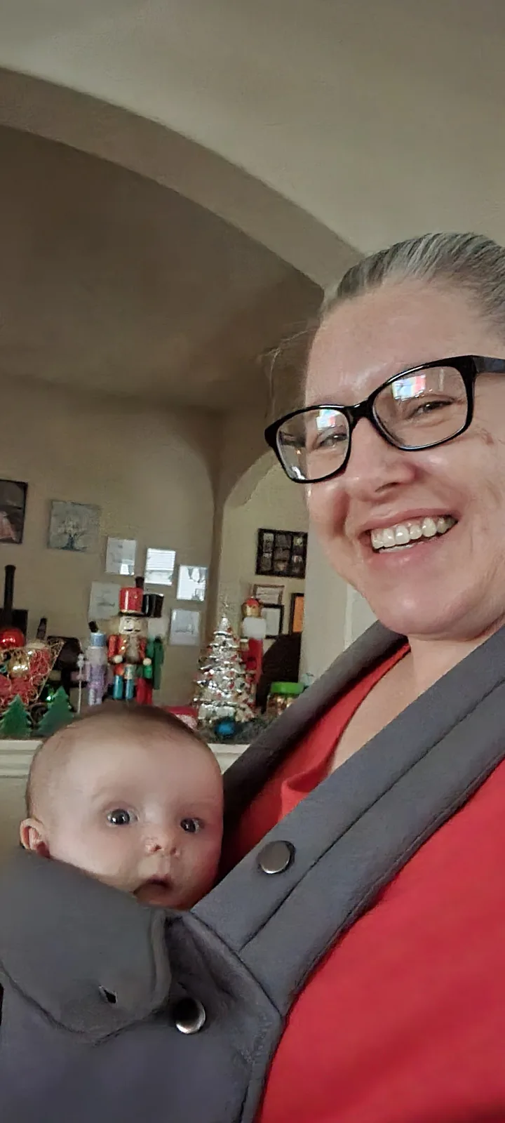 A woman with glasses smiles broadly while holding a baby in a gray carrier; the baby stares blankly ahead with a surprised…