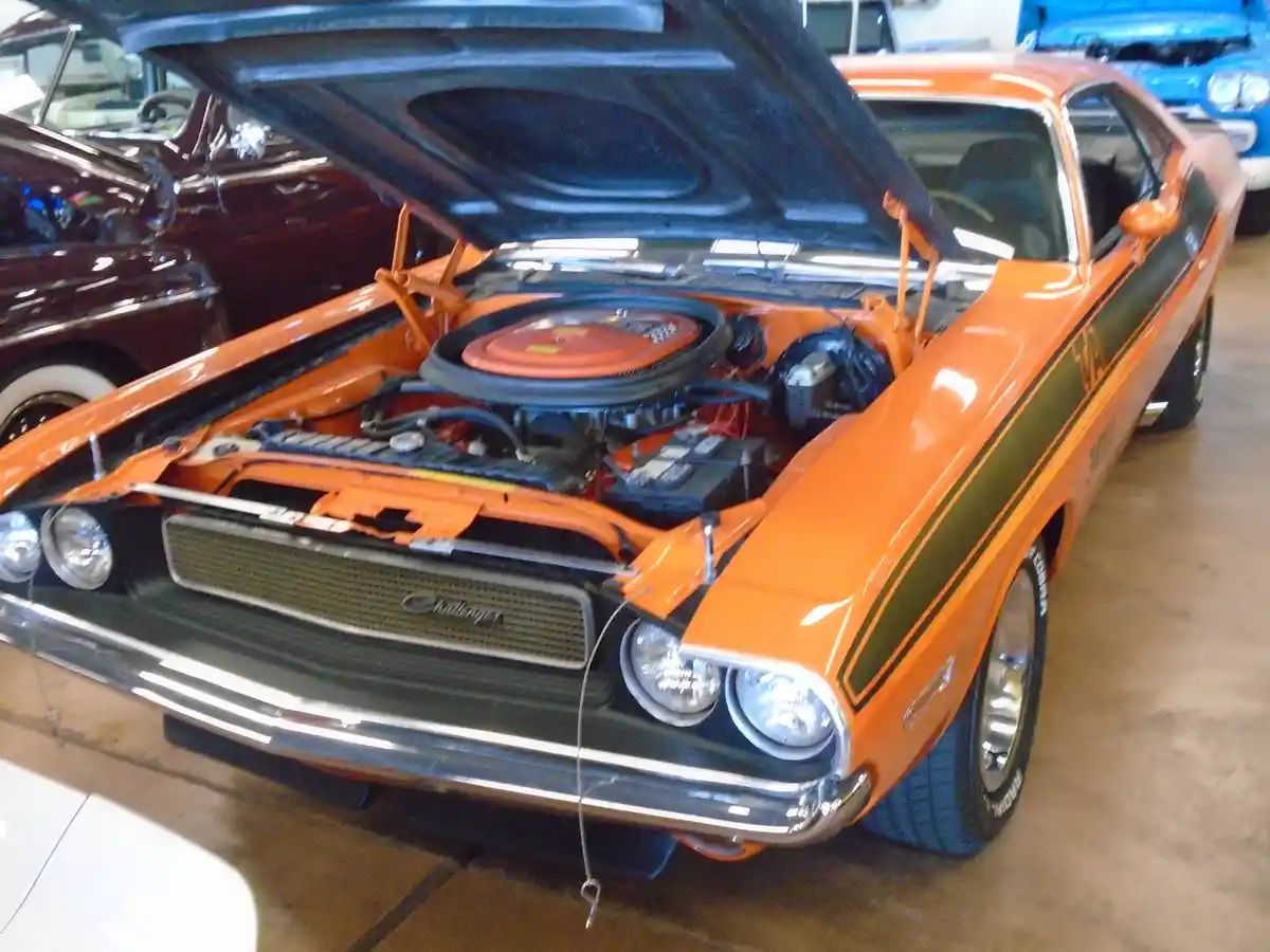 An orange classic Dodge Challenger with its hood open, revealing a detailed engine bay, parked indoors among other vintage…