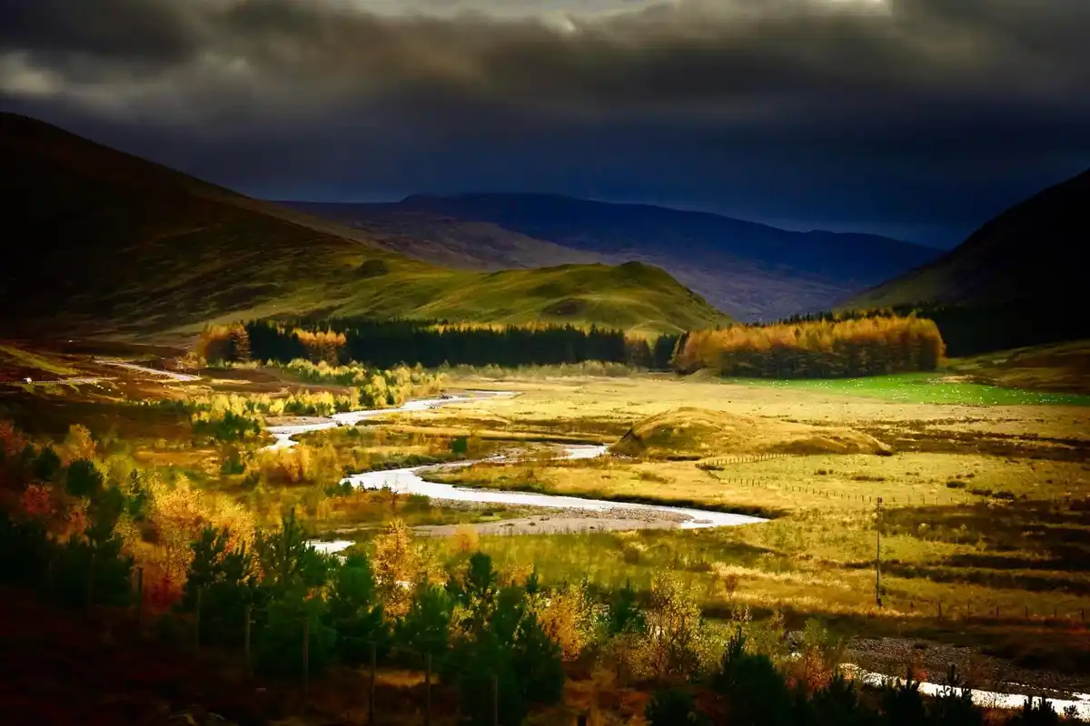 A dramatic landscape photo shows a winding river flowing through a valley with rolling green hills and autumn-colored trees…