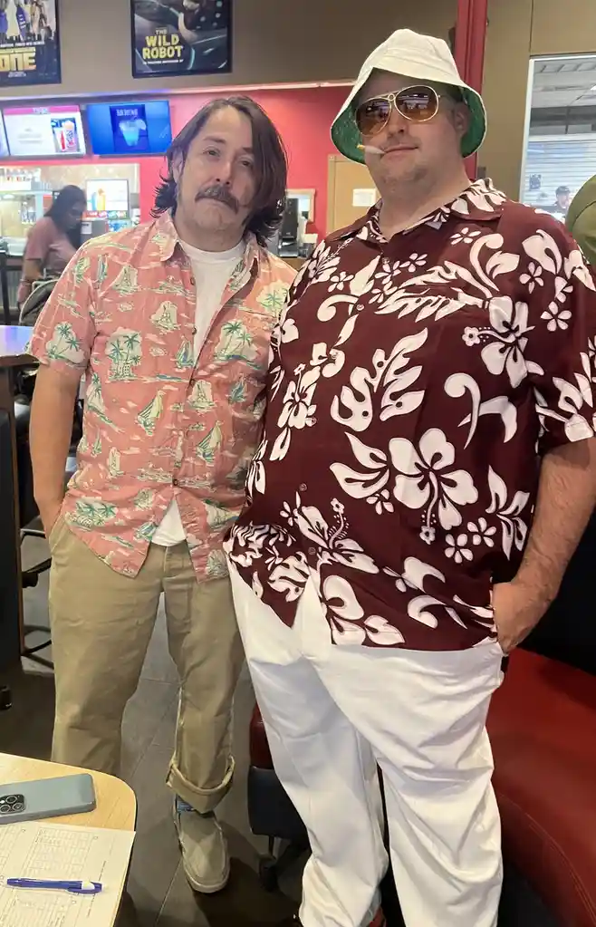 Two men stand side-by-side in a fast-food restaurant, both wearing brightly patterned Hawaiian shirts. The man on the left…