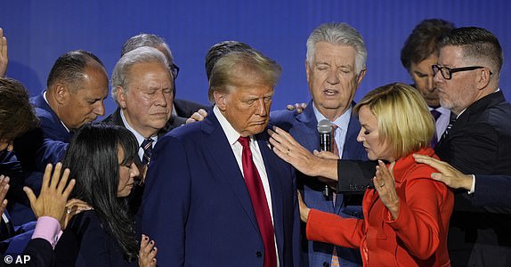 A photo of Donald Trump surrounded by people placing hands on his shoulders and chest, with a woman in a red jacket reaching…
