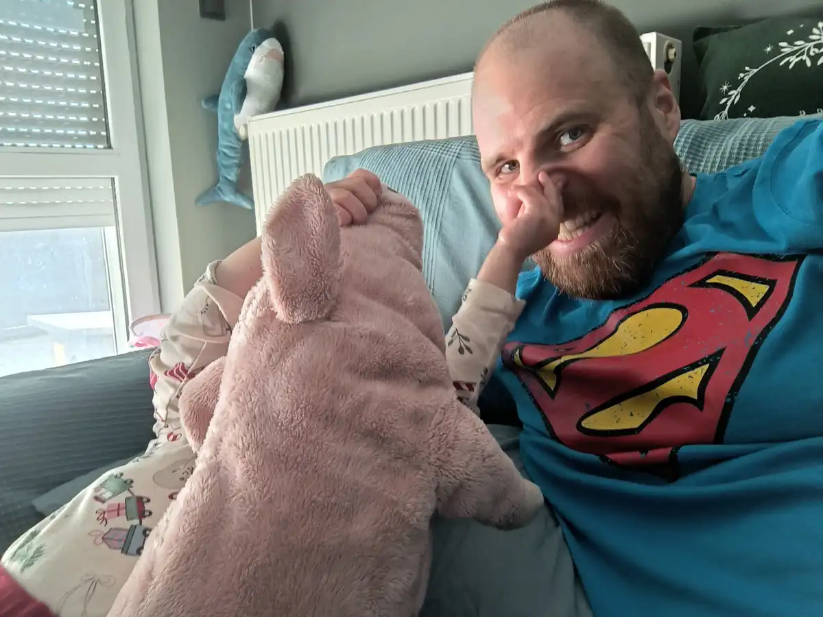 A man with a beard wearing a Superman t-shirt sits on a couch, smiling broadly while a small child in pajamas holds a pink…