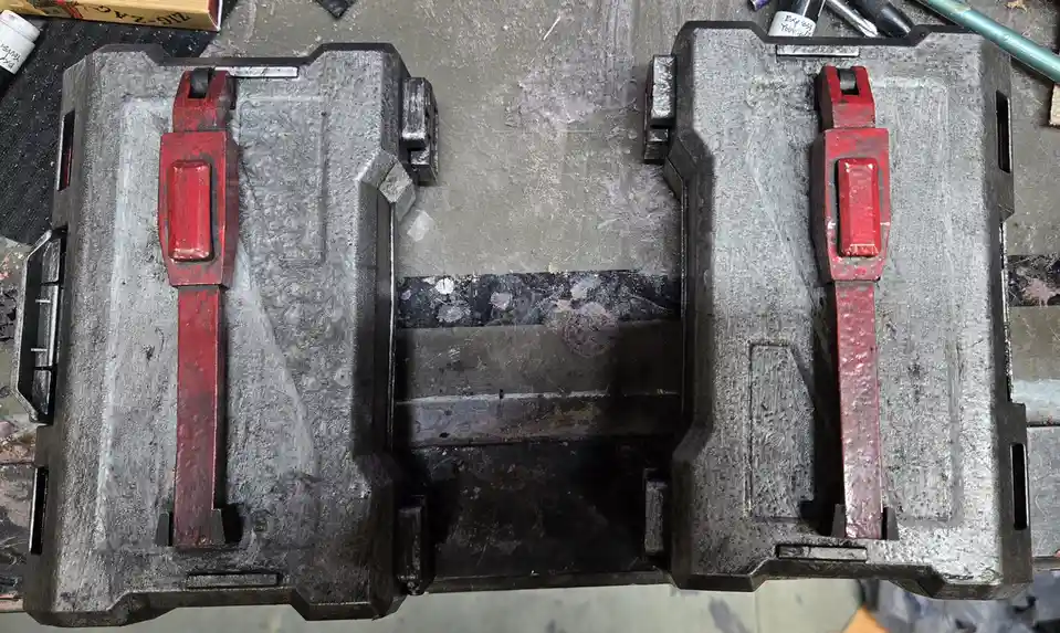 An overhead shot of a heavily used, dirty, gray plastic toolbox with red latches, open to reveal an empty interior. The…