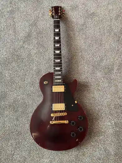 A Gibson Les Paul electric guitar with a cherry sunburst finish lies on a gray carpet. The guitar has gold hardware, two…