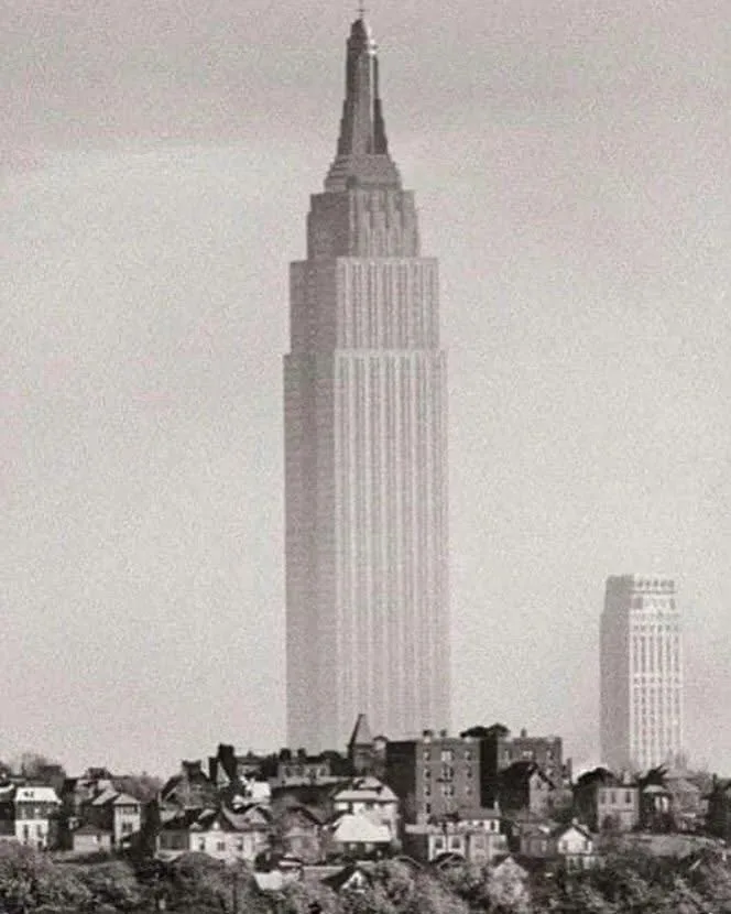 a black and white historical photograph shows the empire state building towering over a cityscape of low-rise buildings…