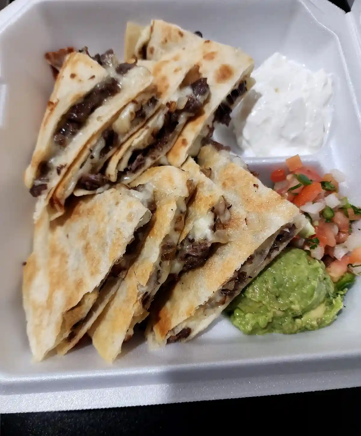 A close-up photo of a white styrofoam takeout container filled with sliced beef and cheese quesadillas, accompanied by…