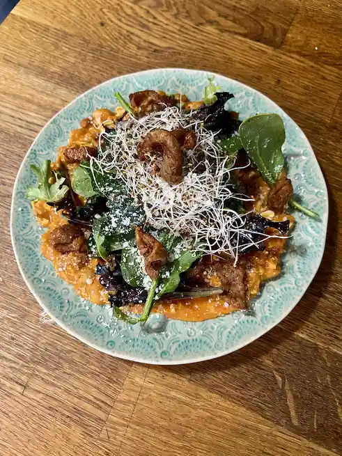 A top-down photo of a plated dish on a wooden table, featuring a base of orange-colored food topped with mixed greens…
