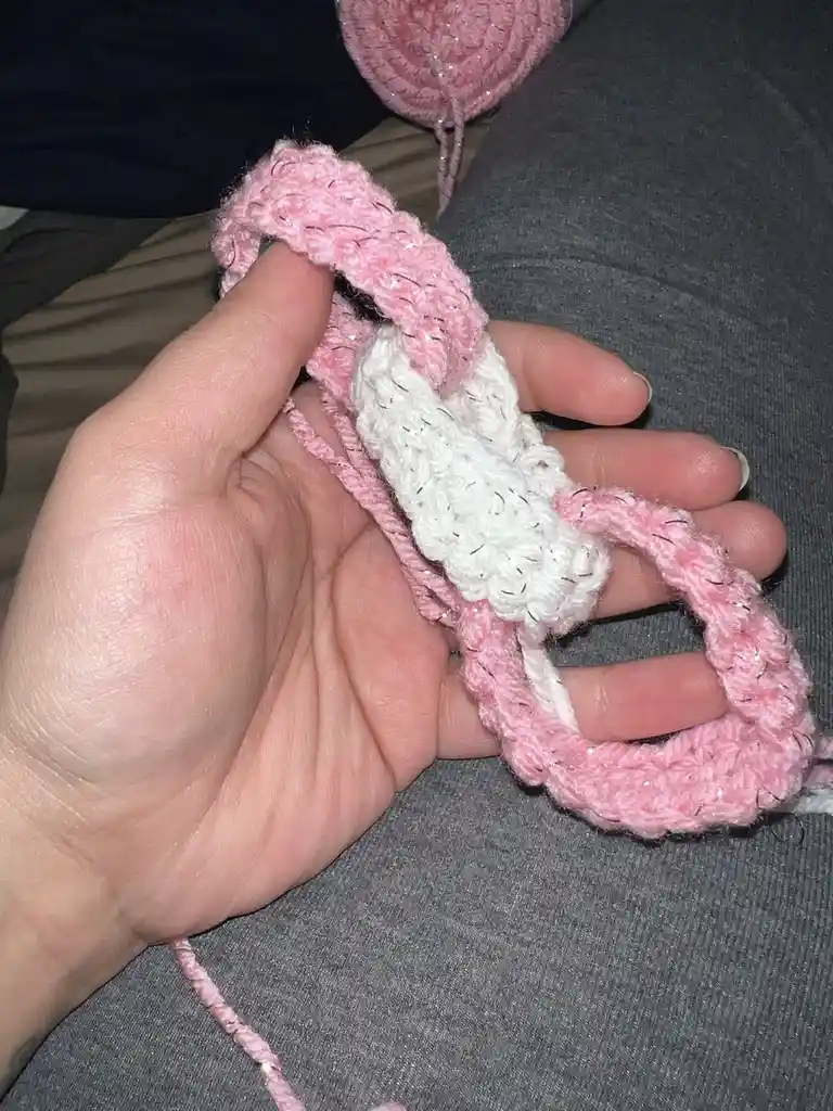 A hand holds a crocheted pink and white yarn piece shaped like a phallus, with a white middle section and pink ends, resting…