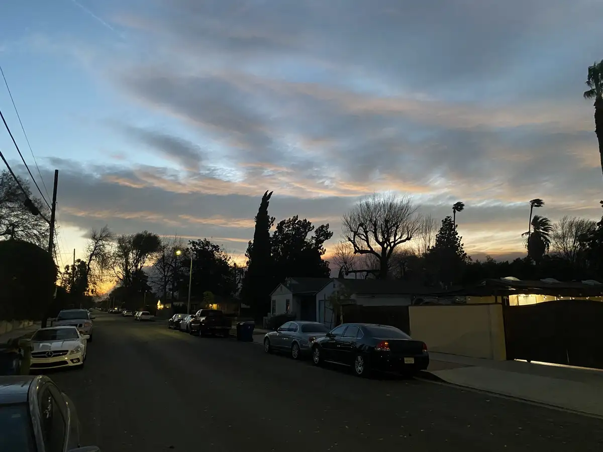 A residential street at dusk with cars parked along both sides, silhouetted trees, and a dramatic sky with orange and blue…