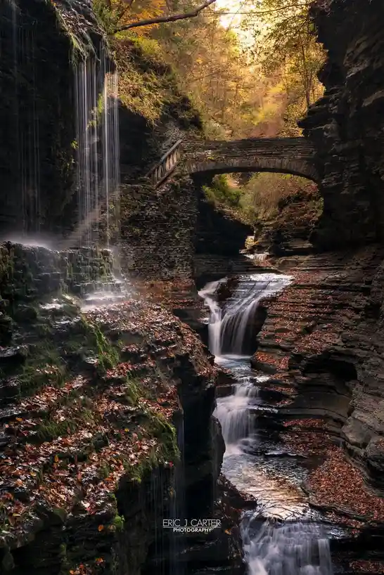 A serene autumn landscape featuring a stone arch bridge spanning a cascading waterfall in a rocky gorge, with moss-covered…