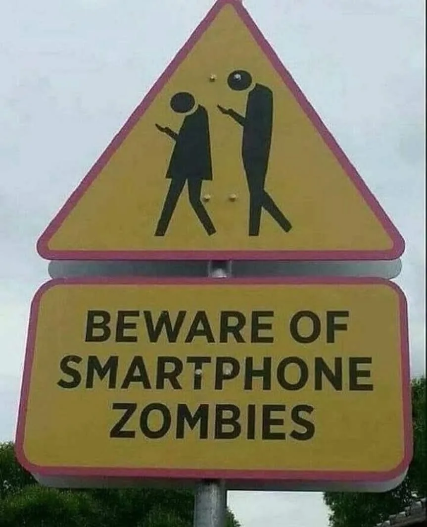 A yellow triangular pedestrian crossing warning sign with two stick figures walking and looking at their phones, overlaid…