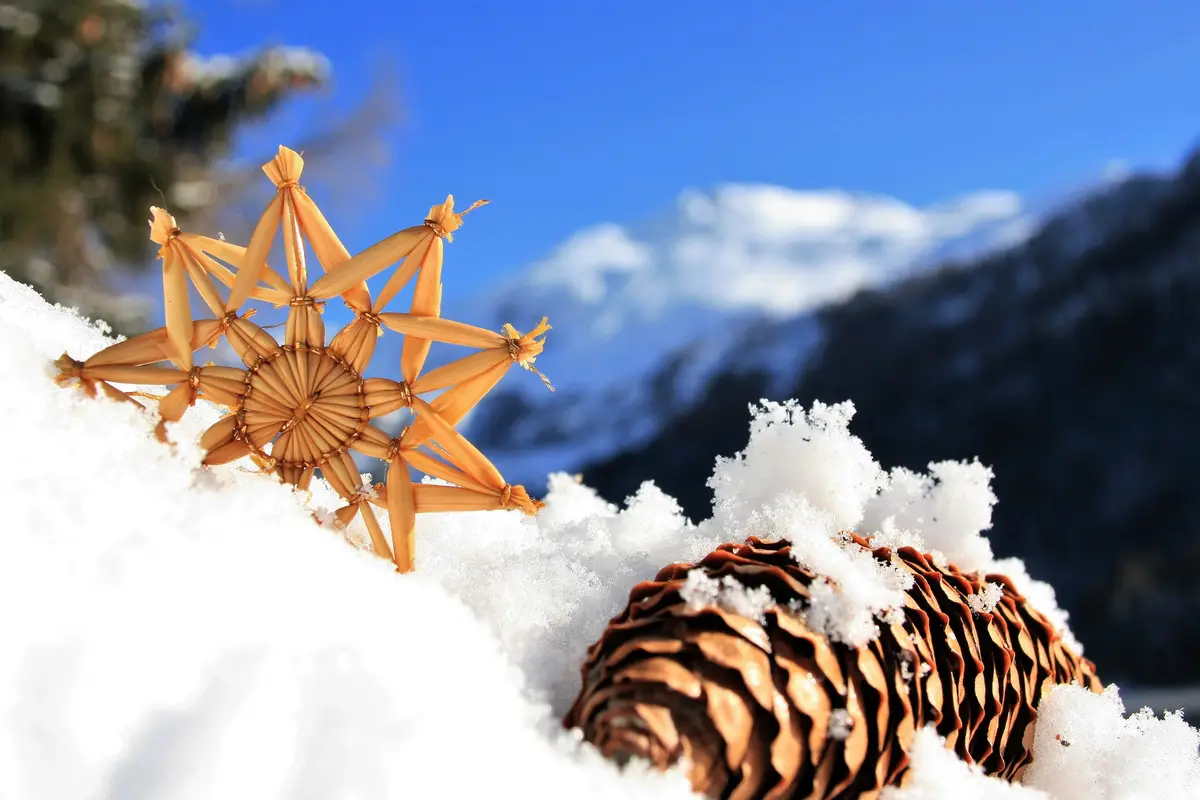 A close-up photo shows a handmade straw star decoration and a pinecone resting in fresh snow, with snow-capped mountains and…