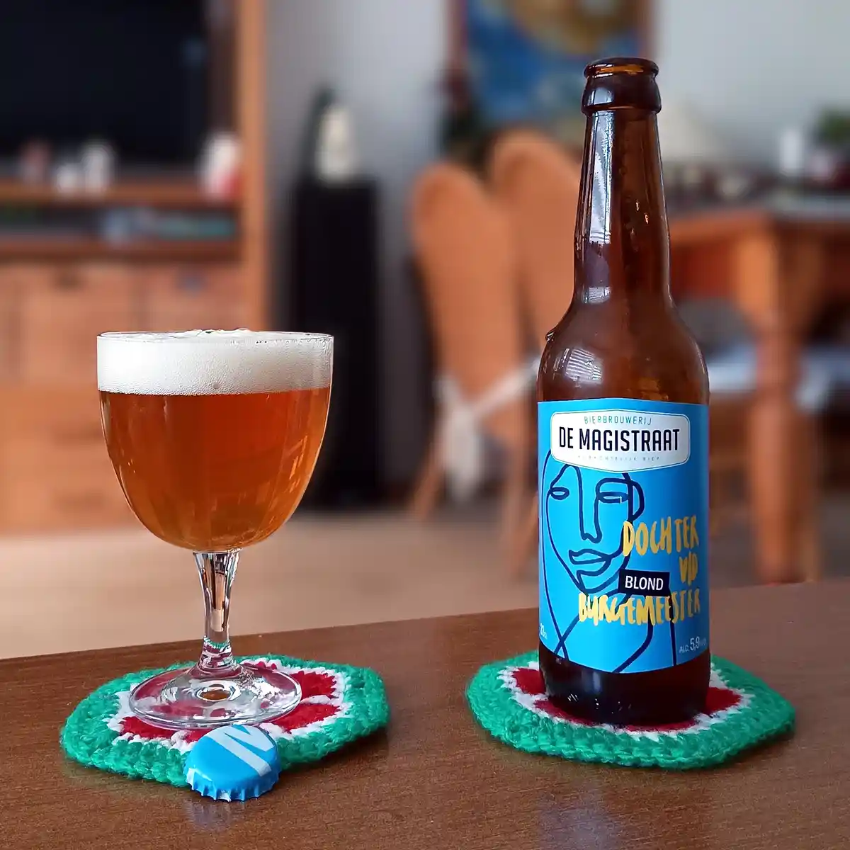 A brown beer bottle with a blue label reading 'De Magistraat' and 'Dochter Vijf Burgemeester' sits next to a filled beer…
