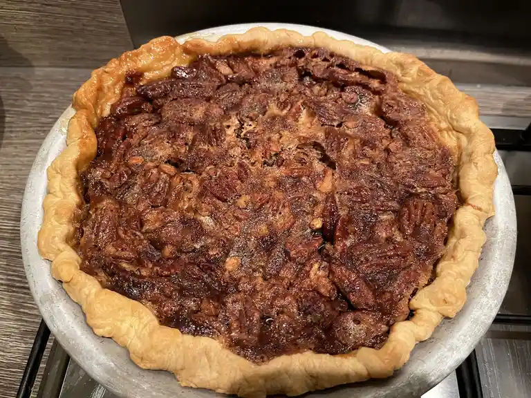A close-up photo shows a freshly baked pecan pie in a metal pie tin, featuring a golden-brown crust and a glossy…