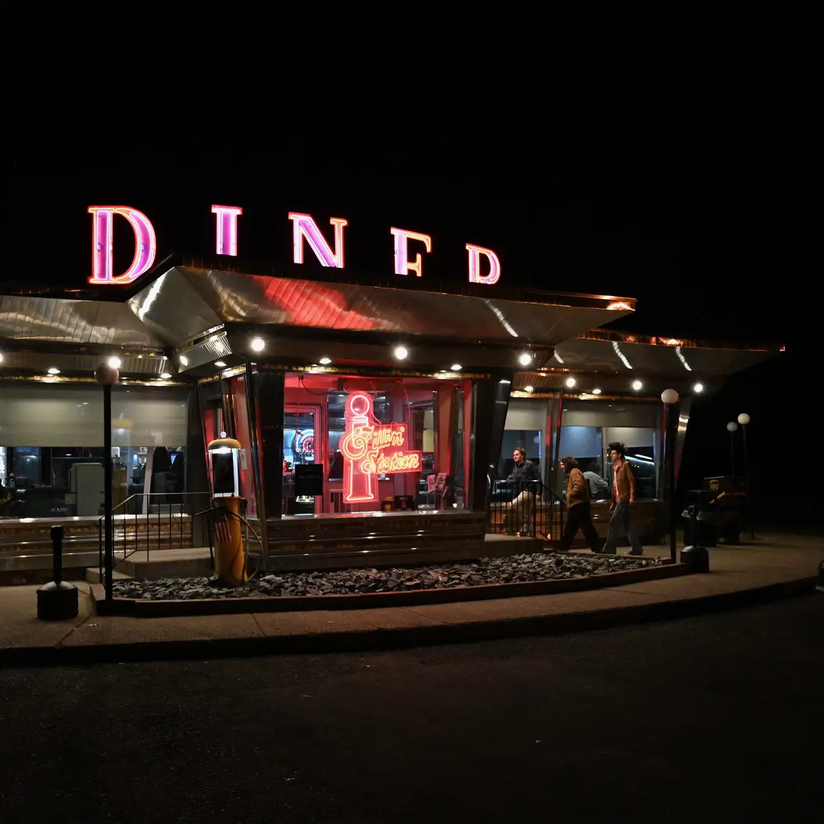 A nighttime photo of a retro-style diner with a large, glowing pink neon sign that reads 'DINFD' above the entrance. Inside…