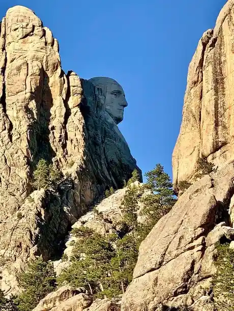 A large, carved stone face resembling a human profile is embedded in a rocky cliffside, framed by two towering rock…