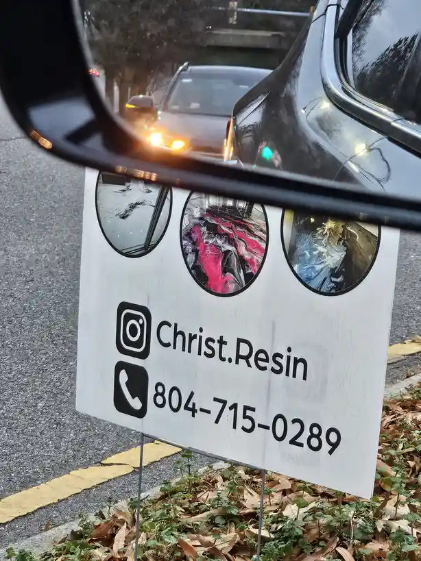 A white business sign for 'Christ.Resin' with an Instagram handle and phone number is seen through a car's side-view mirror…