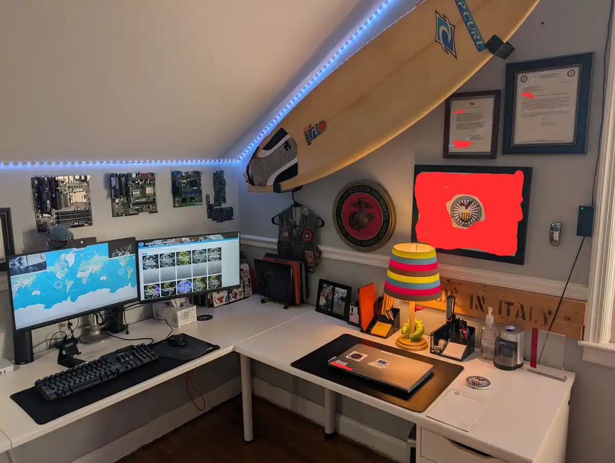 A home office with two monitors displaying satellite imagery, a surfboard mounted on the wall, framed certificates, a Marine…