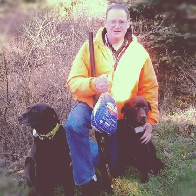 A man in a yellow hunting jacket and glasses sits outdoors with two black Labrador retrievers. He holds a shotgun and a…