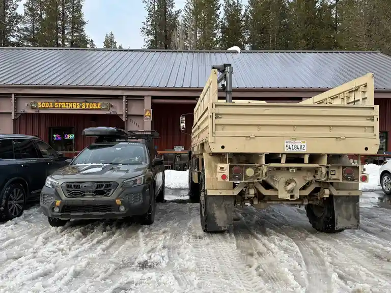 A military-style tan truck with a California license plate is parked in deep snow next to a dark gray Subaru SUV in front of…