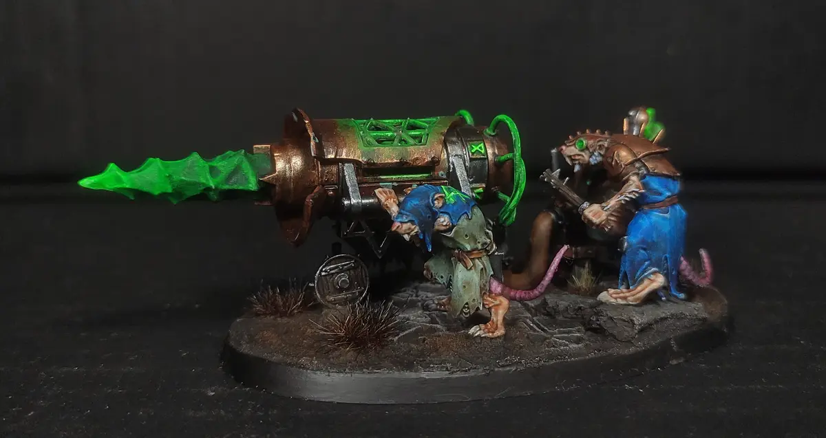 Two painted rat-like miniature figures operate a large, rusted, green-energy-emitting cannon on a rocky base. One rat is…