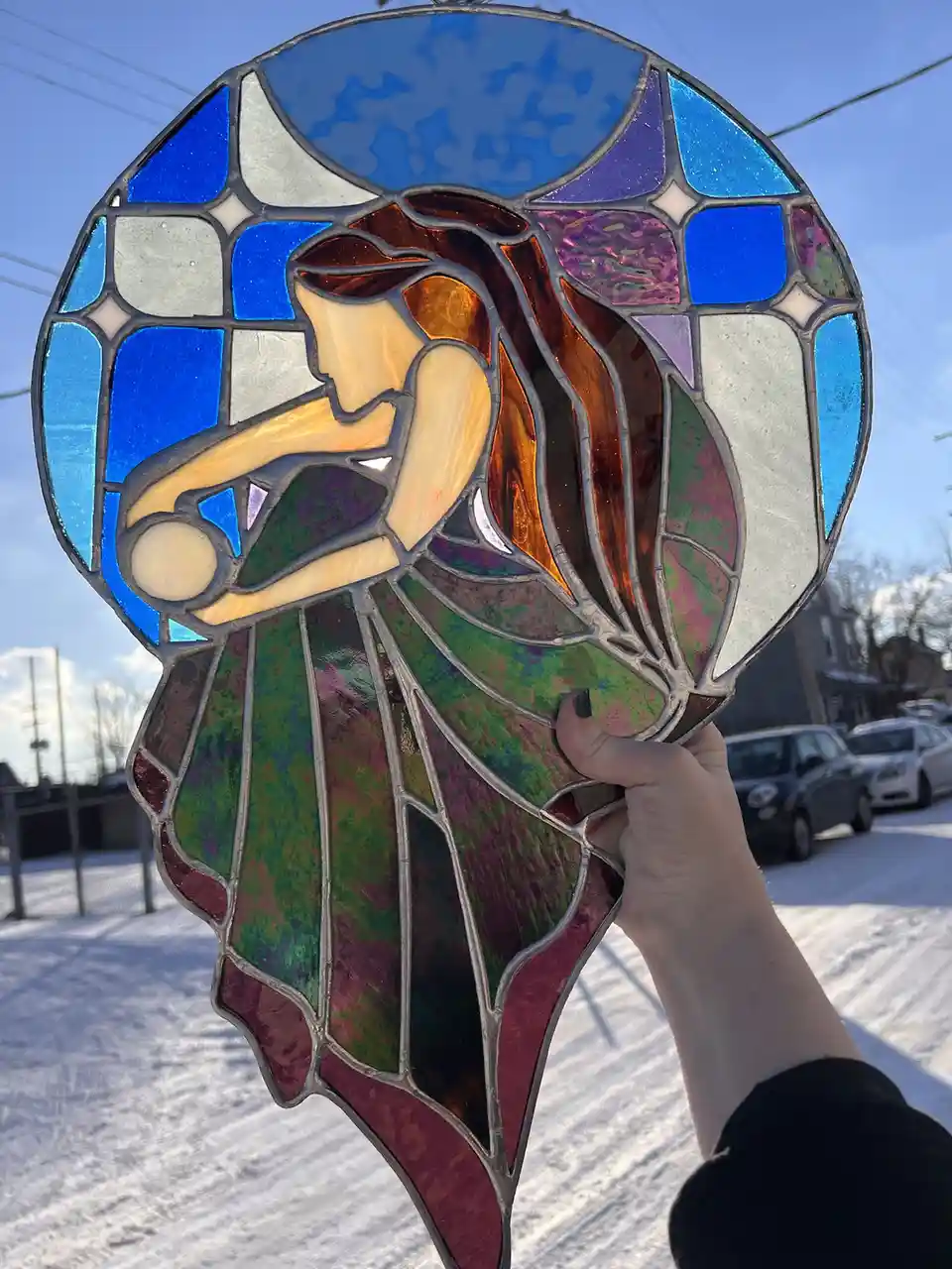 A hand holds up a circular stained glass artwork depicting a stylized female figure with flowing hair and a dress, set…