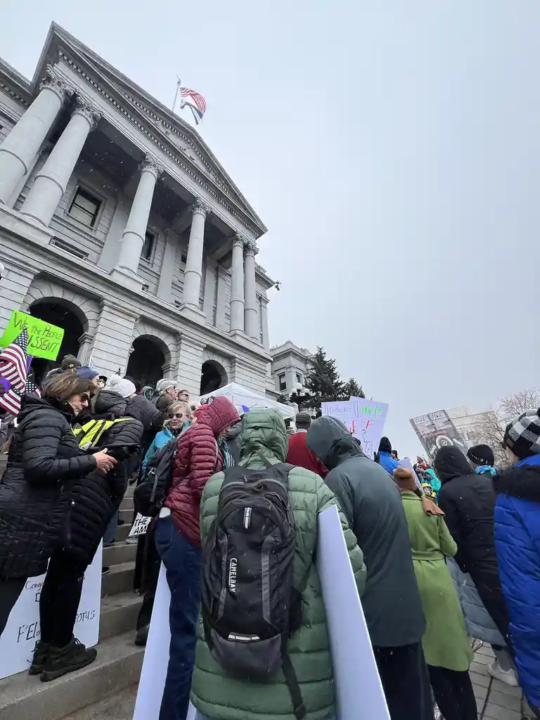 A crowd of protesters in winter coats stands on the steps of a large classical-style government building with columns and an…