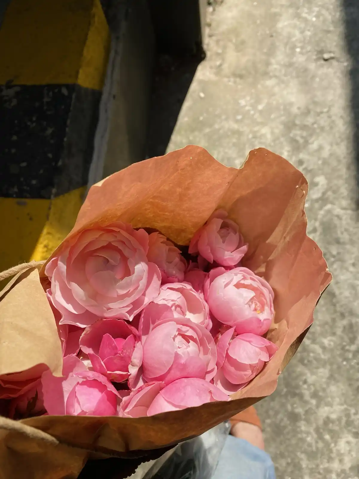 A close-up photo shows a hand holding a bouquet of pink roses and peonies wrapped in brown paper, with a yellow-and-black…
