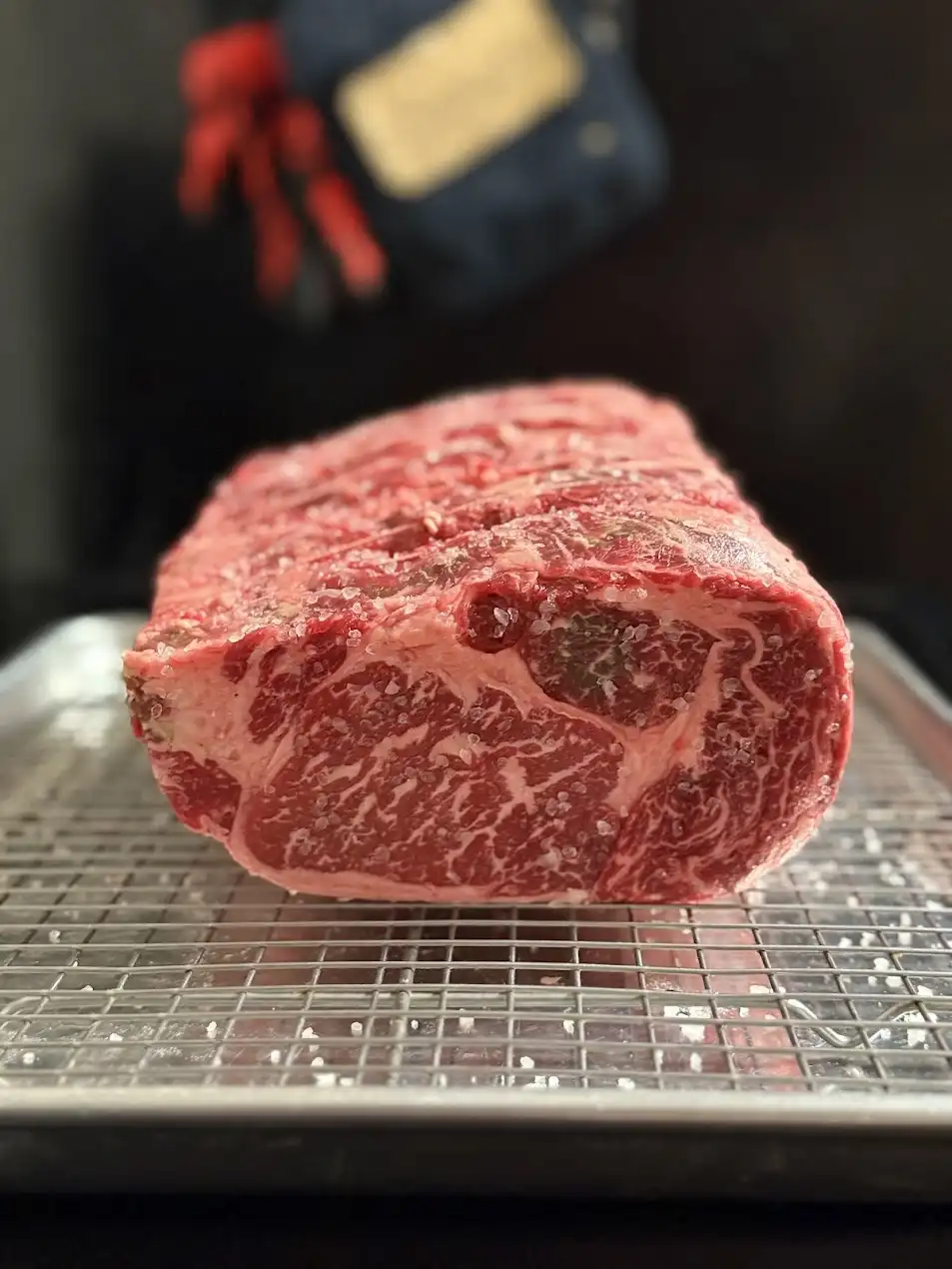 A large, raw, marbled beef roast covered in coarse salt sits on a metal wire rack over a baking sheet. In the blurred…