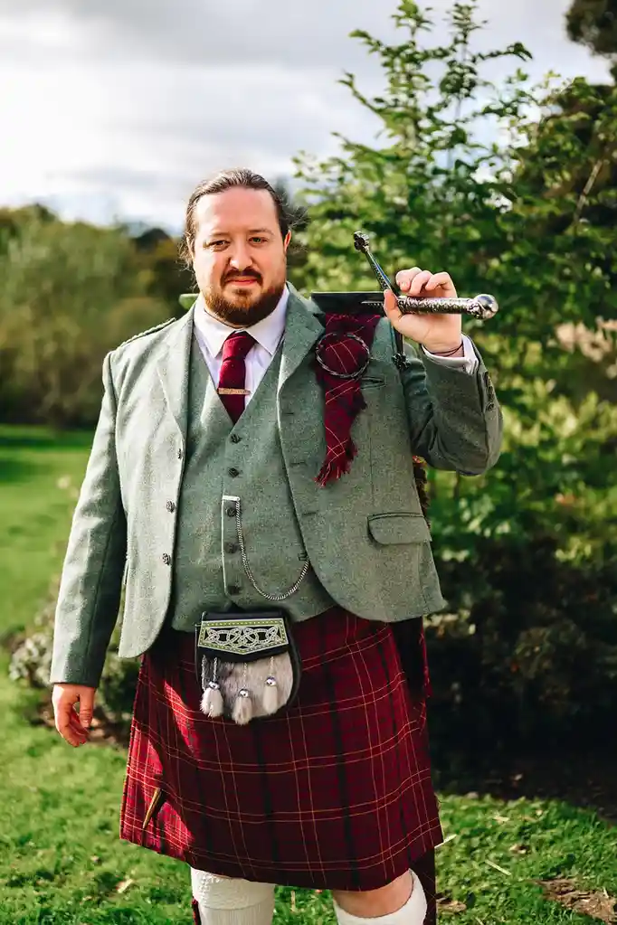 a man in traditional Scottish attire, including a green jacket, vest, red tartan kilt, and sporran, stands outdoors holding…