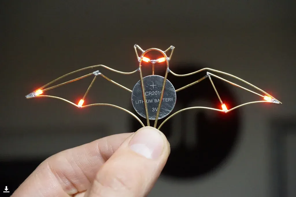 A hand holds a small, intricate bat-shaped object made of thin wire with five glowing orange LEDs at the wingtips and two at…