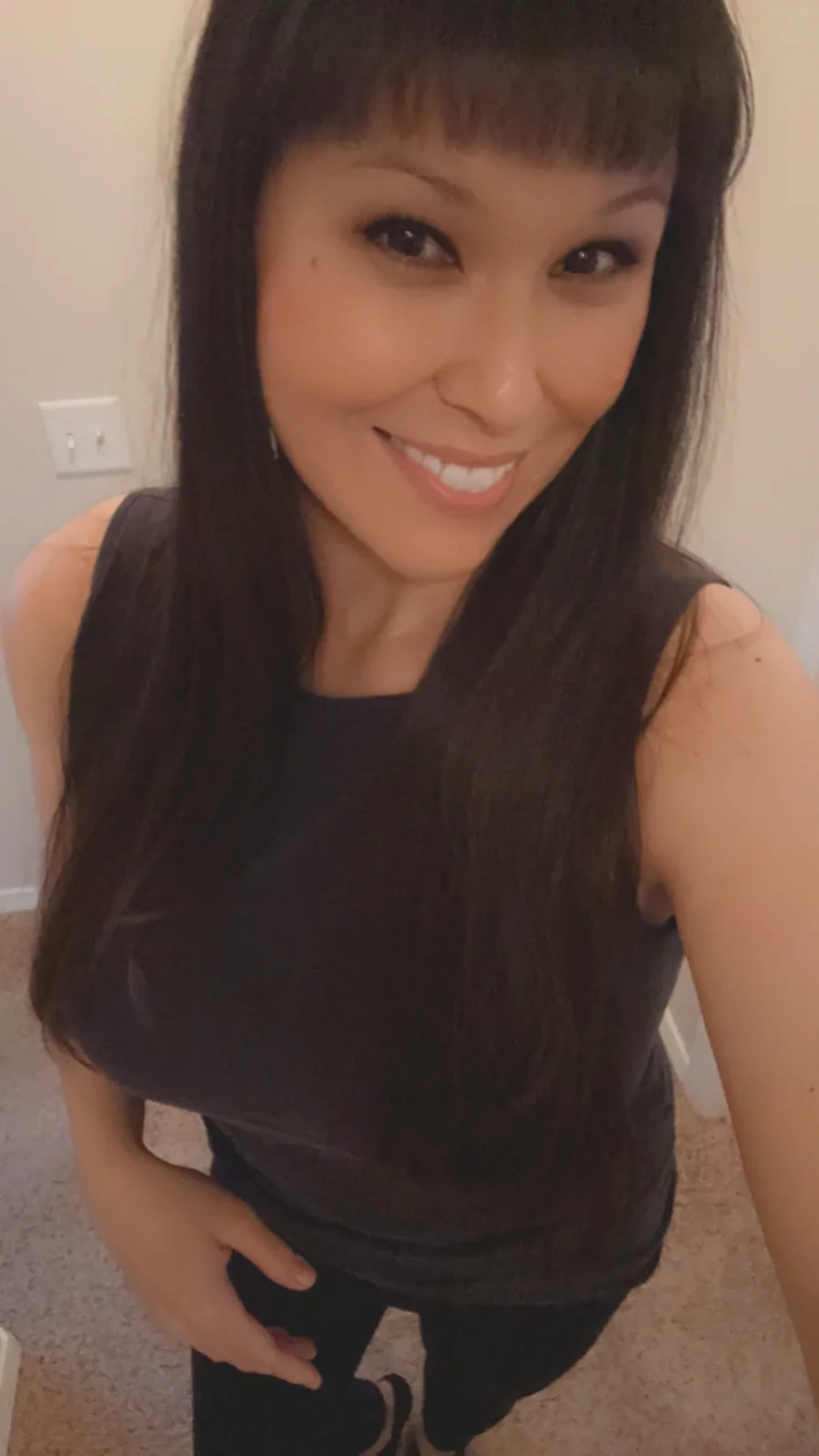 A woman with long dark hair and bangs smiles at the camera while wearing a dark tank top and dark pants, standing indoors on…