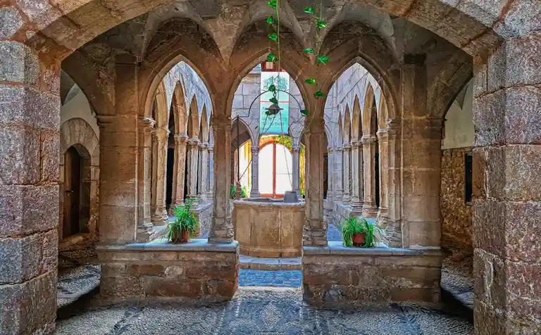 A symmetrical stone cloister with pointed arches and columns, featuring a central fountain, potted plants on stone ledges…