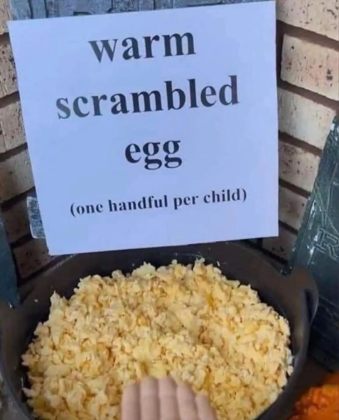 A hand reaches into a cast iron skillet filled with yellow scrambled eggs, with a white sign above reading 'warm scrambled…