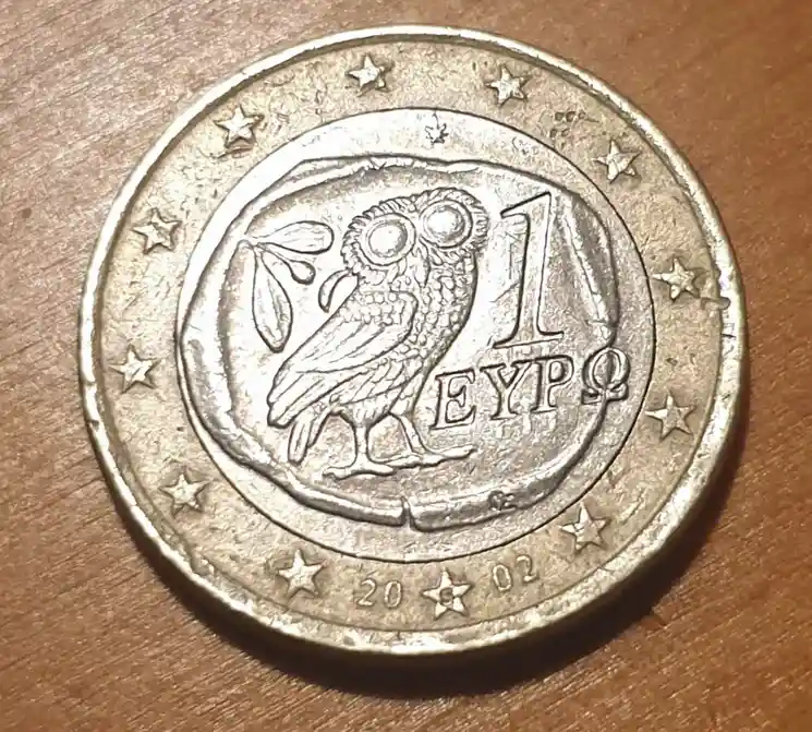 A close-up photo of a 2002 Greek 1 Euro coin, showing its national side with an owl, olive branch, and the denomination '1…