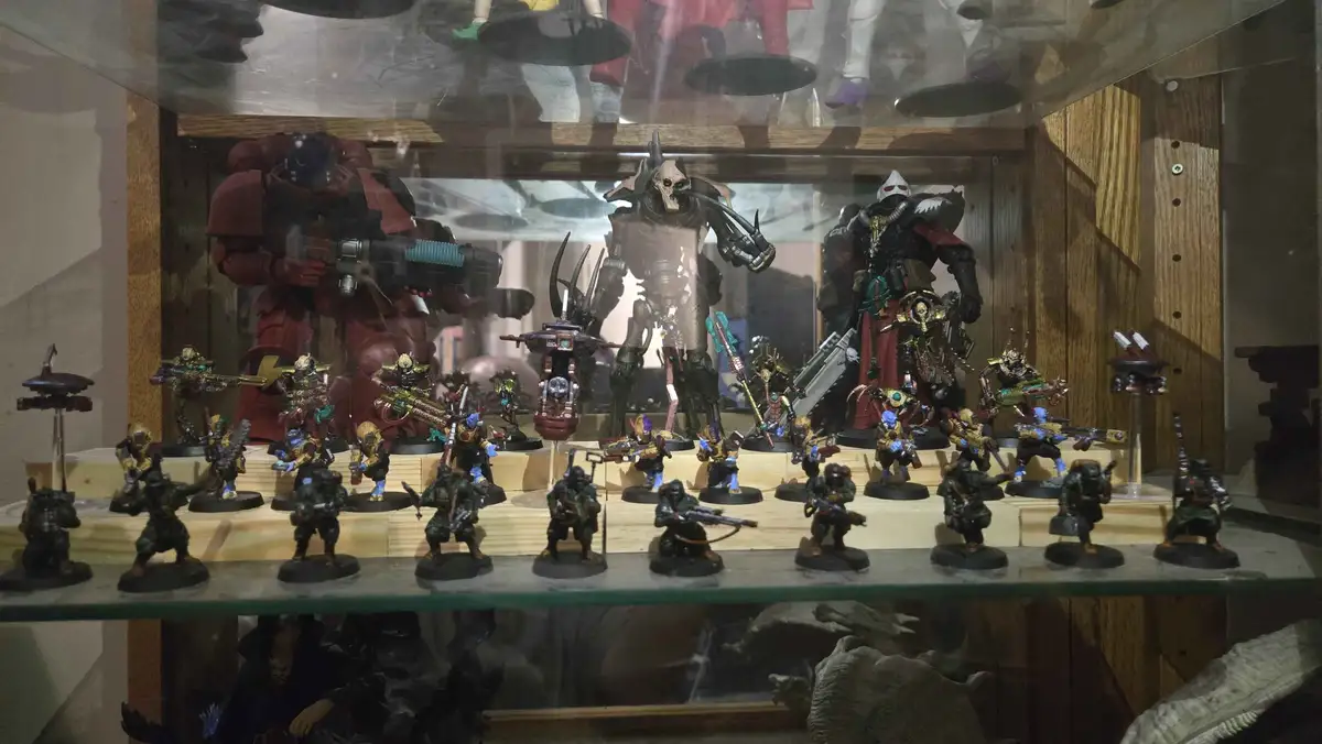 A glass display case holds numerous painted tabletop miniatures, mostly in dark, gritty sci-fi armor, arranged in rows…