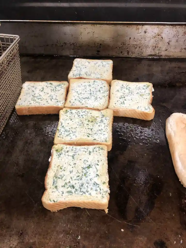 Six slices of buttered bread with green herbs are arranged in a cross shape on a dark, greasy griddle. A metal basket is…