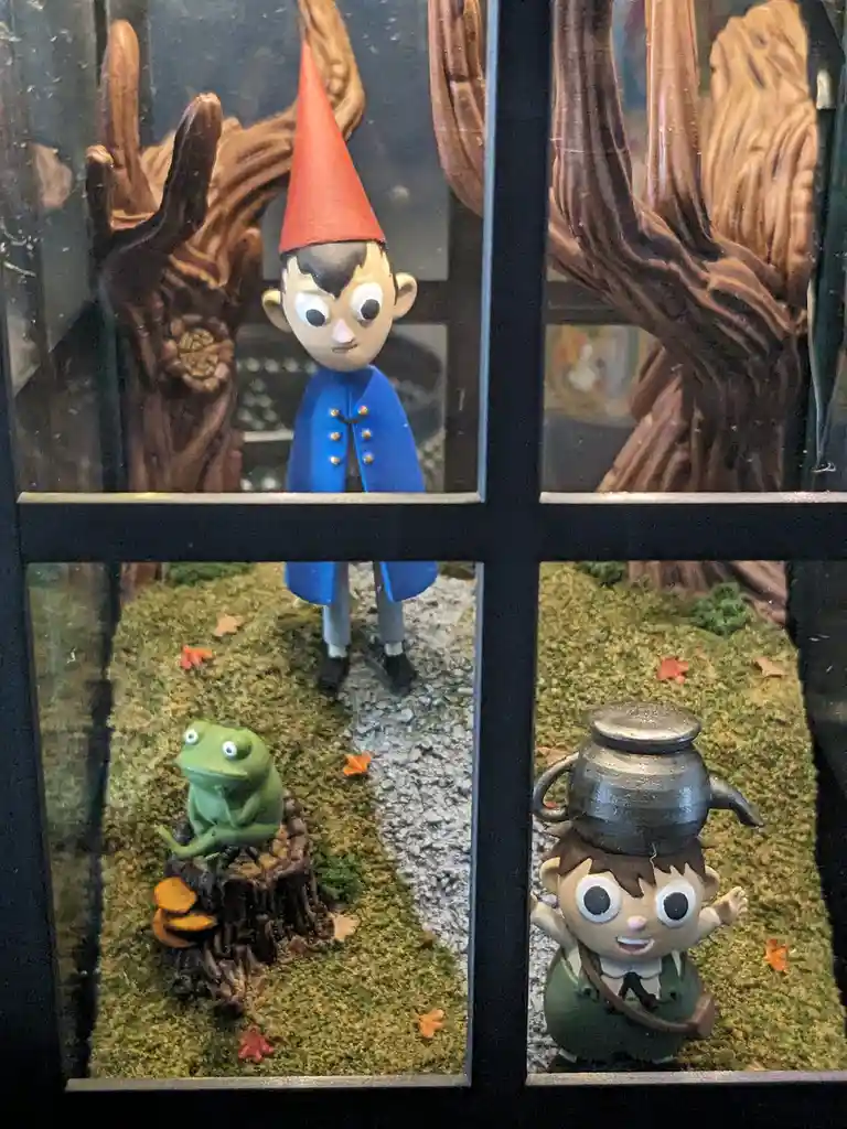 A diorama-style display features three clay-like figurines in a windowpane-divided scene: a boy in a red cone hat and blue…