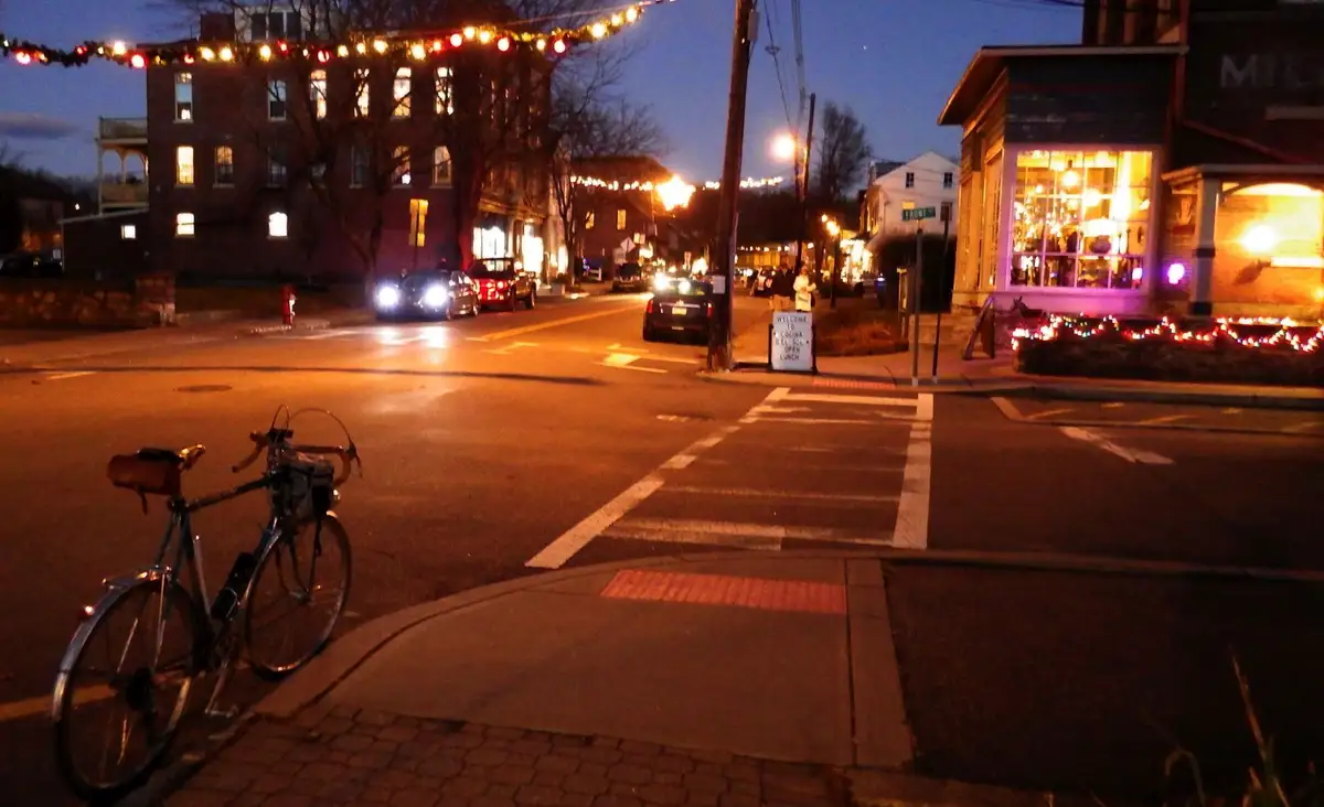 A bicycle is parked on a sidewalk at a street corner during twilight, with festive string lights strung across the street…