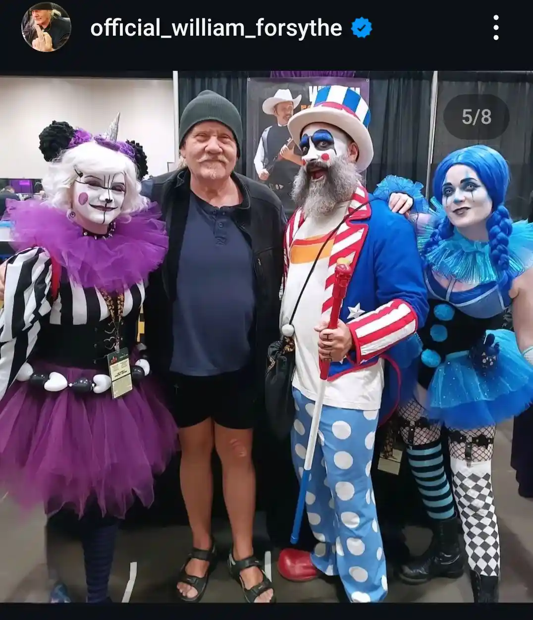 A man in a plain black jacket and beanie stands between two elaborately dressed clowns at a convention. The clown on his…