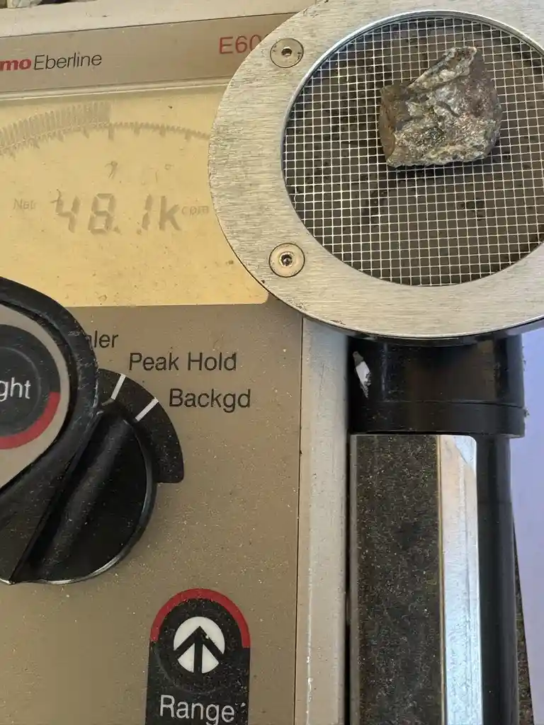 A close-up of a Geiger counter, branded 'Eberline', with a rough, irregular rock resting on its metal grid sensor. The…