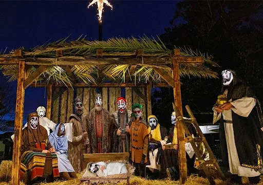 A nighttime outdoor nativity scene with figures in costumes, many wearing white face masks with black eye holes, resembling…