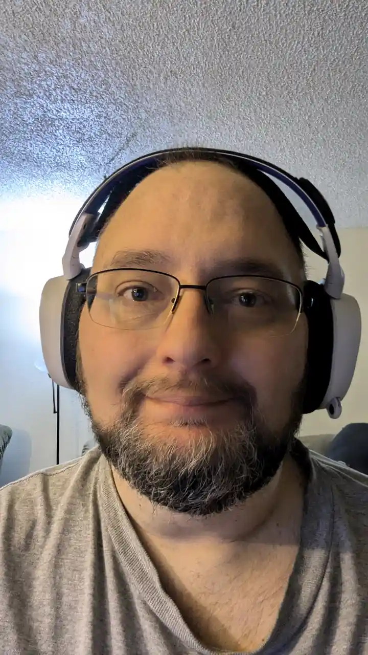 A close-up selfie of a man with glasses and a graying beard wearing large over-ear headphones, looking directly at the…