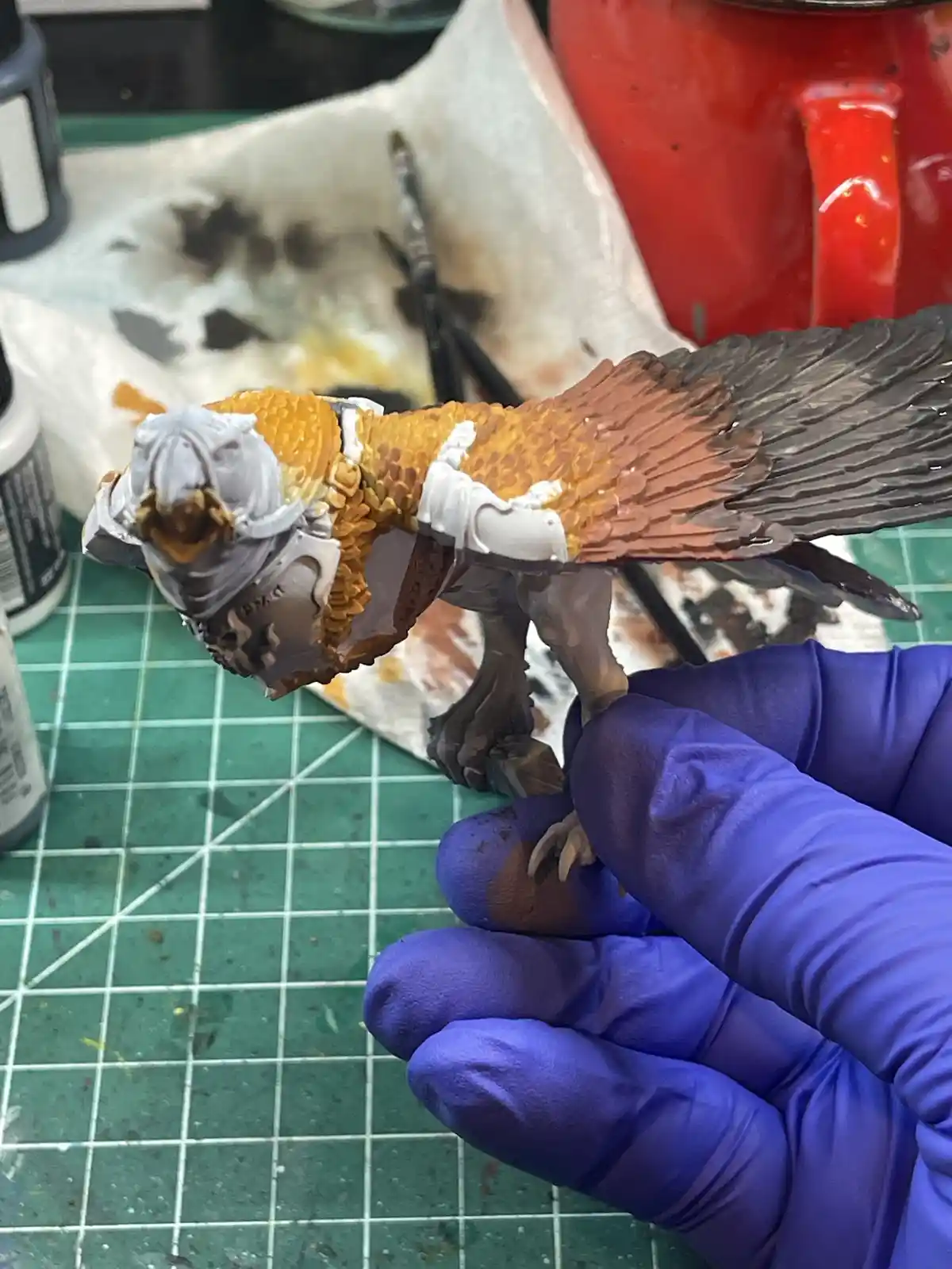 A person wearing blue nitrile gloves holds a painted miniature bird figurine, which has a detailed silver helmet on its…