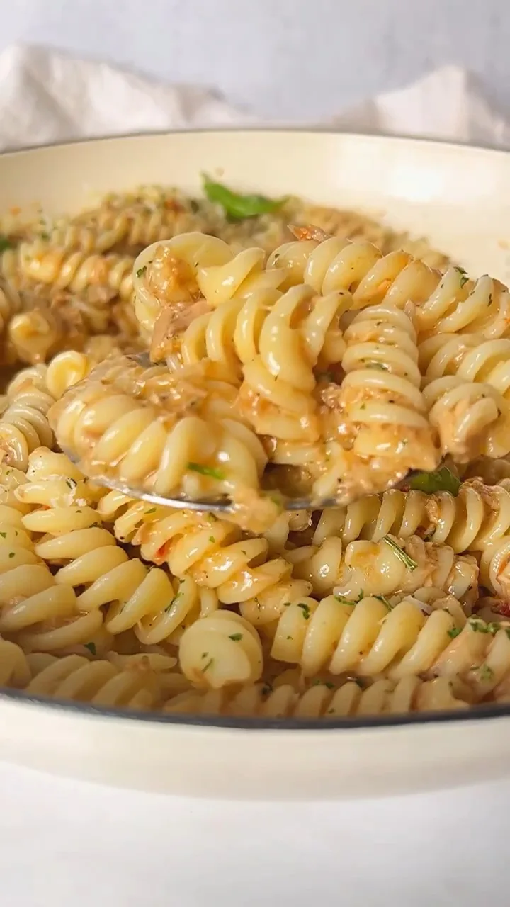 a close-up photo of a spoon lifting a generous portion of creamy, sauce-coated fusilli pasta from a white bowl, garnished…