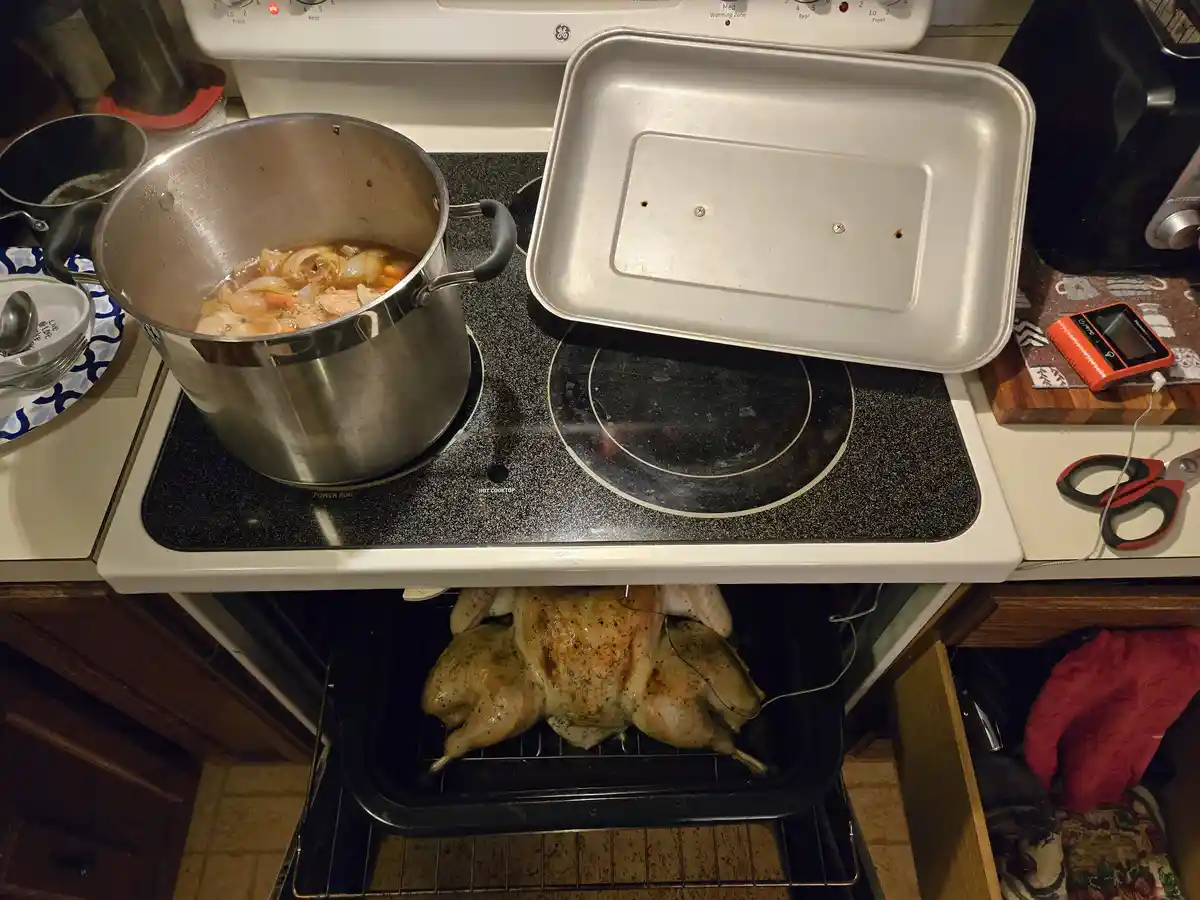 An overhead shot of a kitchen stove with a pot of soup simmering on the left burner and an empty roasting pan lid resting on…